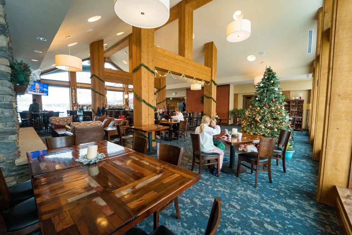 A festive living area featuring a beautifully decorated Christmas tree, exposed wooden beams, and several comfortable seating arrangements. The space is filled with a warm, cheerful glow, accentuated by holiday decorations and a bar area in the background, creating an ambiance of celebration and togetherness.