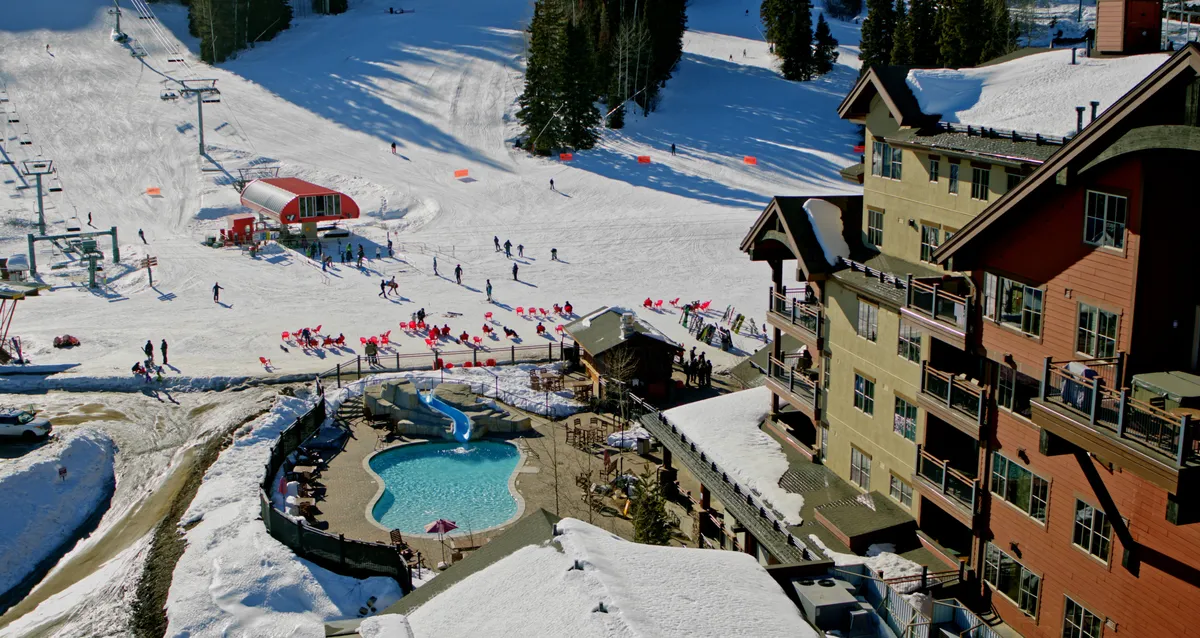 This image captures the exterior pool area adjacent to the hotel, with snow-dusted ski slopes, ski lift, and inviting lounge chairs. The lively scene showcases guests enjoying both poolside relaxation and access to winter recreation, set against the backdrop of dramatic mountains.
