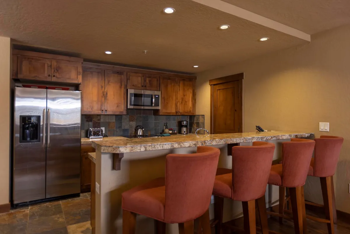 A spacious gourmet kitchen featuring a polished granite countertop with bar stools, gleaming stainless steel appliances, and warm ambient lighting. The kitchen is set up for both cooking and entertaining, with space for social gatherings and hearty meals.