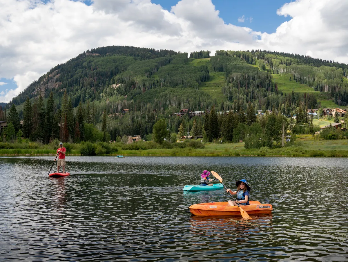 A lively scene featuring people kayaking and paddleboarding on a tranquil lake, with the majestic mountains and lush forest in the background. The image captures outdoor enthusiasts actively engaging with nature, set against the spectacular scenery that defines the area in summer and fall.