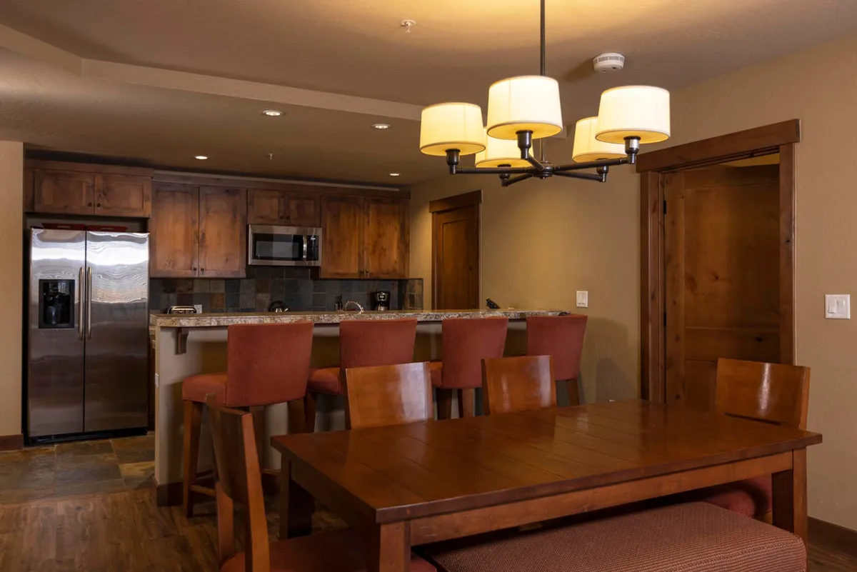 A welcoming interior scene centering on a large dining table adjacent to a kitchen island and lit by a chandelier. The table is set for family meals or gatherings, and the space is arranged for entertaining guests as well as preparing and enjoying meals. The cozy atmosphere is achieved through soft lighting, warm wood tones, and a seamless flow between dining and kitchen areas.