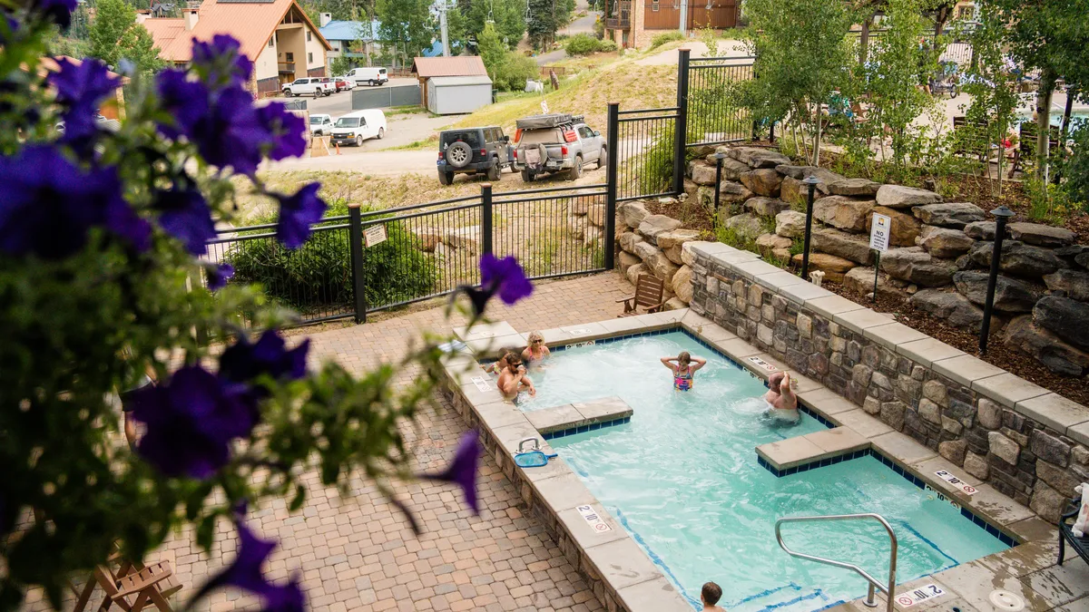 The scene centers on an outdoor hot tub surrounded by lush greenery and flowering plants, with guests unwinding in the water and basking in the sun. The natural setting enhances the sense of relaxation and leisure during warmer months.