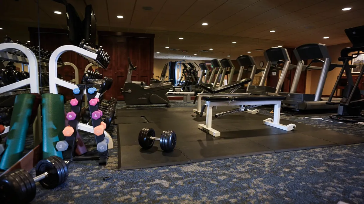 The scene shows a fully equipped fitness center featuring dumbbells, treadmills, a weight bench, and other exercise equipment. The bright, motivating space encourages an active lifestyle and offers opportunities for both solo workouts and group fitness sessions.