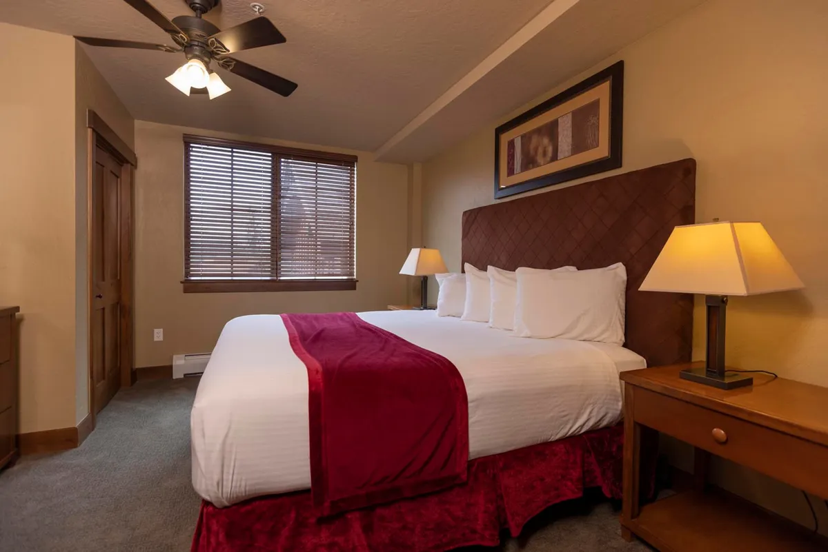 A second inviting bedroom, showcasing a king-sized bed dressed in decorative pillows, a nightstand with a lamp, and natural light streaming through window blinds.