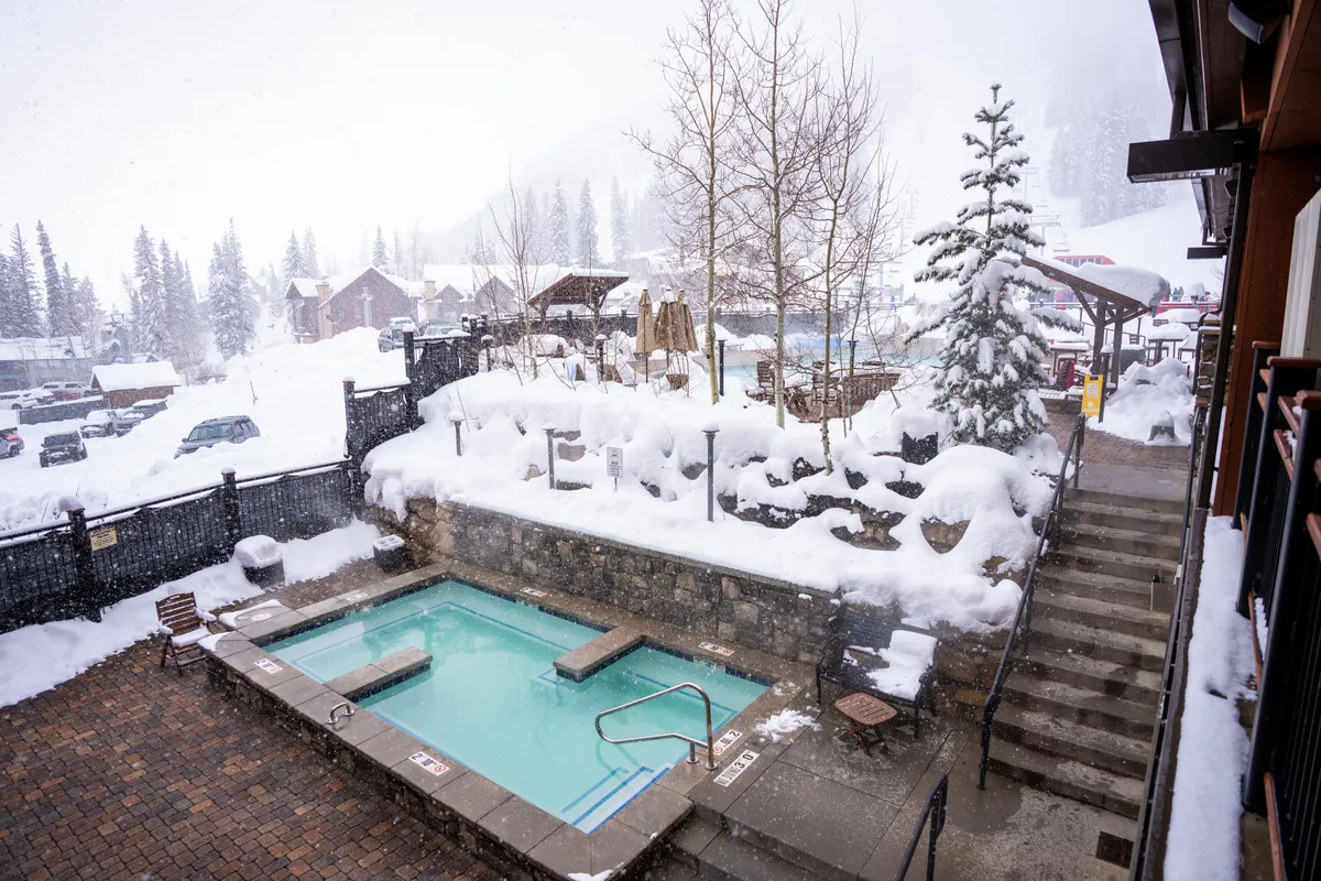 An inviting outdoor hot tub surrounded by snow-laden trees and stone steps, with mountains in the backdrop. Steam rises from the tub as guests relax and soak, enjoying the contrast between the chilly mountain air and warm water. The scene suggests après-ski relaxation and a sense of community in a tranquil, luxurious setting.