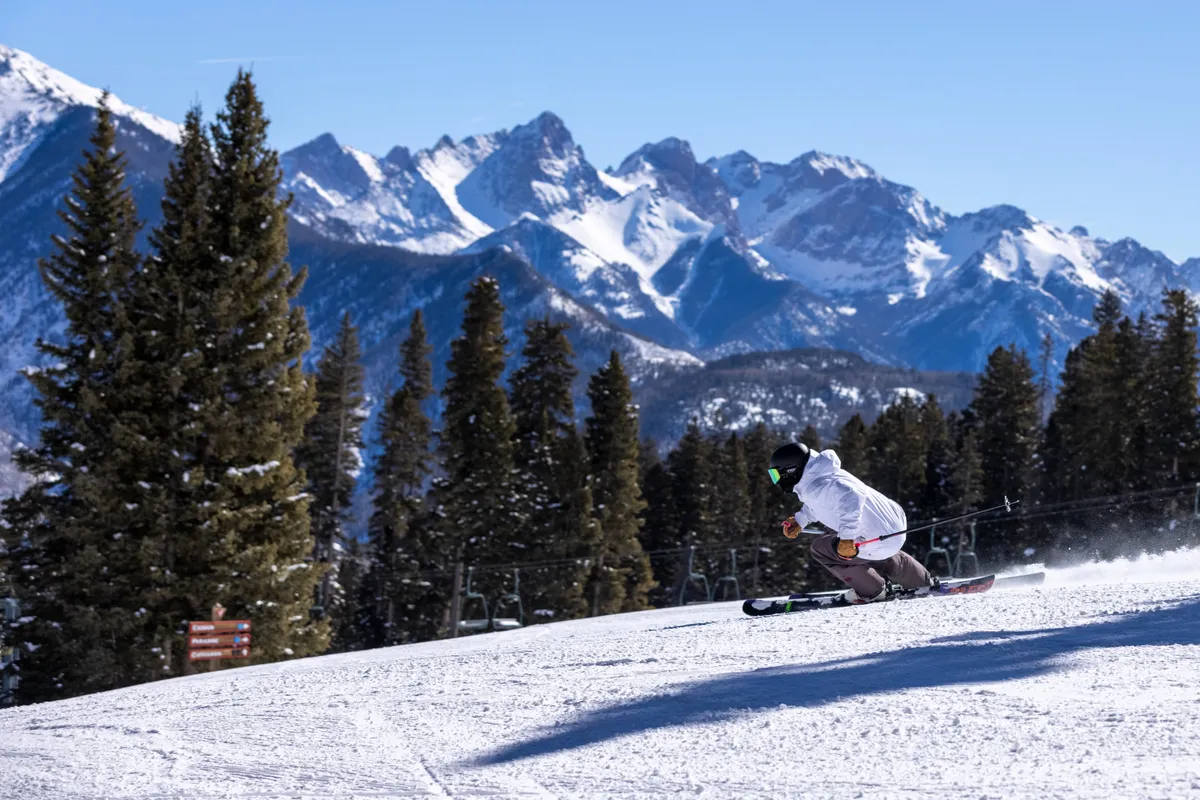 A dynamic action shot of a skier carving down pristine, snow-covered slopes, with a dramatic mountain range stretching into the distance. The bright winter sunlight and expansive white landscape capture the energy and thrill of the mountain experience at Purgatory Resort.