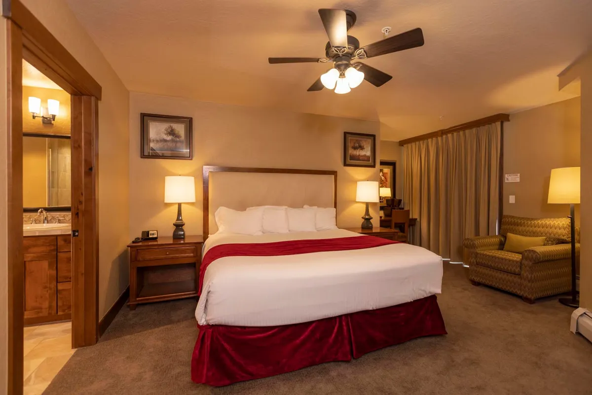 A calm, inviting bedroom featuring a king-sized bed with plush bedding, nightstands topped with lamps, a comfortable sofa chair, and a large window with curtains that lets in soft natural light.