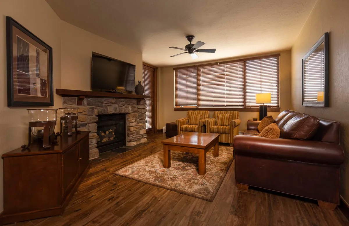 The image showcases a welcoming living room in a luxury ski-in/ski-out condo, centered around a beautiful stone fireplace. The space features a plush leather sofa, comfortable striped armchairs, and a wooden coffee table, all arranged to encourage relaxation and conversation. The warm lighting and cozy furnishings create an inviting, homey atmosphere perfect for unwinding after a day on the slopes.